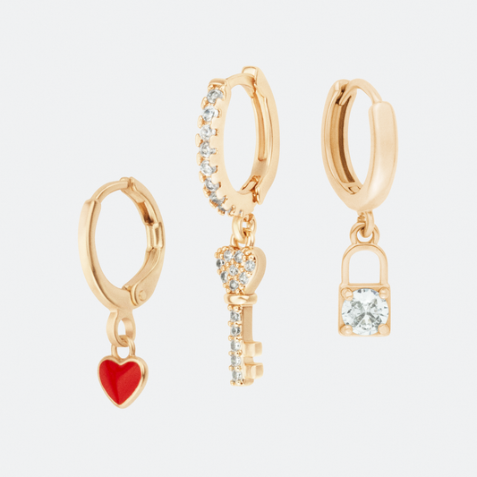 The Amuletum charm hoop earring set by Ocarina, featuring three 18K gold-plated hoops with symbolic dangling charms: a red enamel heart, a zircon-encrusted key, and a sparkling padlock. A meaningful and elegant jewelry collection representing love, mystery, and protection, displayed against a clean white background.