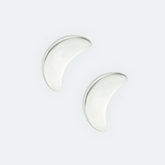 The Lumen earrings by Ocarina, minimalist silver crescent moon-shaped studs with a smooth, polished finish. A celestial-inspired jewelry piece symbolizing light and transformation, displayed against a clean white background with a small blue accent.